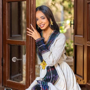 Puede incluir: Una mujer con un vestido crema con intrincados bordados negros, azules y rojos. El vestido tiene un cuello en V y una broche amarilla en forma de cruz. Se apoya en un marco de puerta de madera, sonriendo a la cámara.