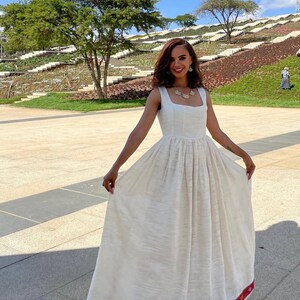 May include: A long, white dress with a square neckline and a full skirt. The hem of the dress features a red and gold patterned border. The woman is smiling and wearing a necklace. The background includes trees and a landscaped area.