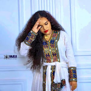 Puede incluir: Un vestido blanco con intrincados bordados en azul, amarillo y rojo. El vestido presenta mangas largas con puños detallados y un panel bordado a juego en la parte delantera. Un collar dorado y una faja blanca completan el look.