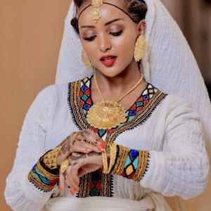 Puede incluir: Una mujer con un vestido blanco tradicional con bordados coloridos alrededor del cuello y los puños. Lleva joyas de oro, incluyendo pendientes, un collar, pulseras y una diadema. El vestido se complementa con un velo blanco.