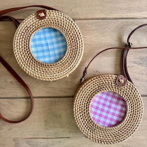 Può includere: Due borse a tracolla rotonde in rattan intrecciato con cinturini in pelle marrone. Una borsa presenta una fodera interna in tessuto a quadretti vichy azzurro e bianco, mentre l'altra ha una fodera in tessuto a quadretti vichy rosa e bianco. Entrambe le borse hanno una piccola linguetta in pelle marrone al centro.