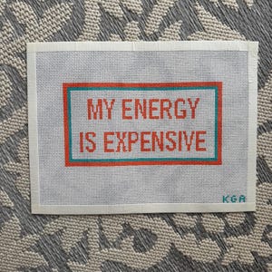 May include: A white needlepoint canvas with the text "MY ENERGY IS EXPENSIVE" in orange letters, framed by a teal and orange border. The canvas is on a gray patterned surface.