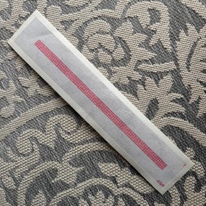 Può includere: Una tela per punto croce rettangolare con un bordo geometrico rosa. La tela è bianca con sfondo grigio e bordo rosa. La tela è su una superficie a motivi grigi e bianchi.