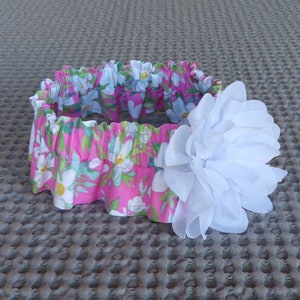 May include: A pink and white floral headband with a large white fabric flower.
