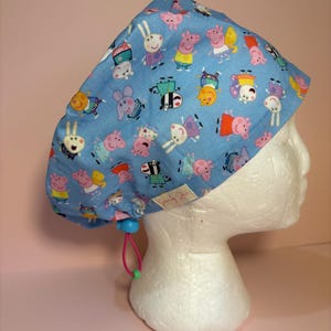 Può includere: Una cuffia chirurgica azzurra con un motivo ripetuto di personaggi di Peppa Pig in vari abiti. La cuffia include un cordino elastico rosa con un fermo blu per la regolazione e una piccola etichetta in tessuto bianco con scritto Sewn by Staci.