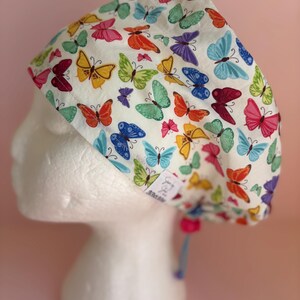 May include: A white scrub cap patterned with a variety of colourful butterflies. The butterflies are in shades of red, orange, yellow, green, blue, and purple. The cap has a pink drawstring and a small tag.