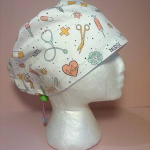 Può includere: Una cuffia chirurgica bianca con un motivo ripetuto di icone mediche tra cui stetoscopi, siringhe, cerotti, forbici e cuori, con la parola NURSE stampata in nero. La cuffia è dotata di un fermo verde e di un cordino di colore rosa.