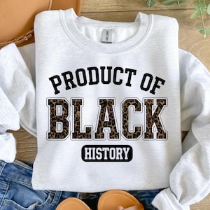 May include: A light gray sweatshirt with the text "PRODUCT OF BLACK HISTORY" in black and leopard print. The sweatshirt is a Gildan brand and is made of a soft material.