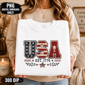 Könnte beinhalten: Ein weißes Sweatshirt mit Rundhalsausschnitt und einem großen Aufdruck der Buchstaben USA in einer strukturierten rot-weiß-blauen Optik. Unter den Buchstaben steht der Text EST. 1776, ergänzt durch einen Stern und Blattmotive.