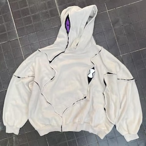 Op de afbeelding: Crèmekleurige hoodie met een uniek ontwerp met zwarte stiksels en een gescheurd stof effect. De capuchon heeft een paars oogdetail en de borst een zwart-witte afbeelding. De hoodie heeft een relaxte pasvorm en lange mouwen.
