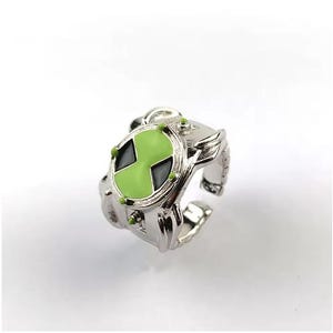 May include: A silver-colored ring featuring a green and black emblem. The ring has a detailed design with small green accents around the emblem. The ring is adjustable and appears to be made of metal.