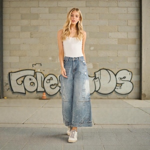 Puede incluir: Una persona con una camiseta sin mangas blanca y vaqueros de mezclilla de pierna ancha y lavado claro con detalles desgastados. Los vaqueros tienen un diseño de patchwork único. Lleva zapatillas blancas. El fondo presenta un muro de ladrillo con graffiti.