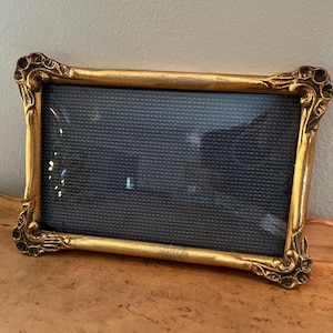 May include: A rectangular gold-colored picture frame with ornate detailing on the corners. The frame holds a dark blue or black textured background. The frame is resting on a light brown surface.