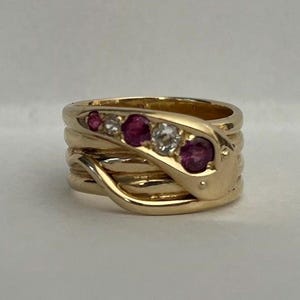 May include: A gold-toned snake ring with a coiled design. The ring features three red gemstones and two clear gemstones set along the snake's head. The ring is set against a neutral background.