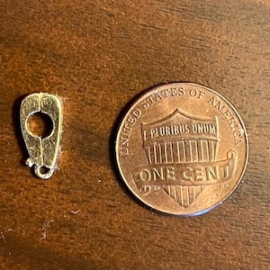 May include: A small, gold-colored jewelry clasp with a circular opening and a loop at the bottom, next to a United States one-cent coin. The clasp is on a brown wooden surface.