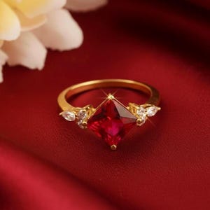 Peut inclure: Bague en or avec une pierre rouge carrée au centre. La bague est ornée de petites pierres blanches de chaque côté de la pierre principale. Le tout est posé sur un fond rouge.