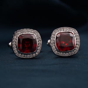 May include: A pair of silver cufflinks featuring a square red gemstone set within a halo of smaller white gemstones.