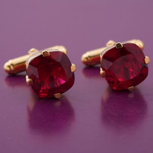 Può includere: Un paio di gemelli dorati con grandi pietre preziose rosso rubino quadrate. Le pietre sono sfaccettate, catturando la luce. I gemelli hanno un design classico, adatti per occasioni formali.