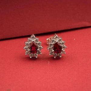 Könnte beinhalten: Ein Paar Ohrstecker mit einem tropfenförmigen, tiefroten Edelstein, umgeben von einem Kranz aus kleineren, klaren Steinen. Die Ohrringe sind in einem silberfarbenen Metall gefasst und liegen auf rotem Grund.