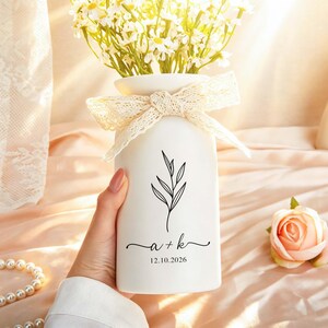 May include: White ceramic vase with a black leaf design and the initials "a + k" with the date "12.10.2026". The vase is adorned with a lace bow and filled with white flowers.