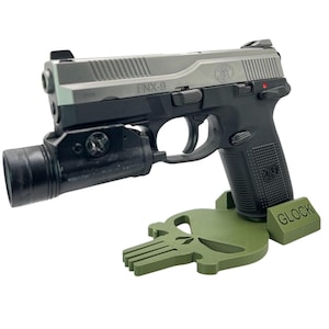 Puede incluir: Una pistola FNX-9 plateada y negra con una luz táctica negra unida al cañón. La pistola está apoyada en un soporte de calavera de Punisher verde impreso en 3D con la palabra "GLOCK" impresa en el lateral.