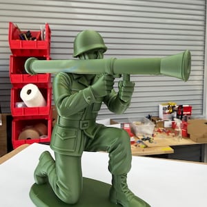 Pode incluir: Uma estatueta de plástico verde-oliva fosco de um soldado em posição de joelhos, usando capacete e uniforme. O soldado segura um grande lançador de foguetes tipo bazuca sobre os ombros, com ambas as mãos segurando o punho da arma.
