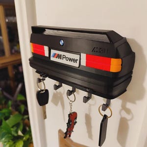 Puede incluir: Llavero negro con forma de parte trasera de coche, con luces traseras rojas, blancas y naranjas. Presenta el logotipo de BMW y la marca "M Power". Varias llaves cuelgan de los ganchos de abajo. Un artículo decorativo para los entusiastas de los coches.