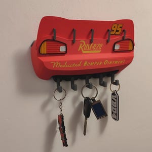 May include: A red key holder shaped like the back of a race car, with the number 95 and the word "Rusteze" in yellow. It has several hooks for keys and the text "Medicated Bumper Ointment".