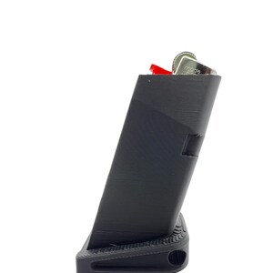 May include: A black plastic magazine holder with a red lighter inside. The holder has a textured surface and a small hole on the bottom.