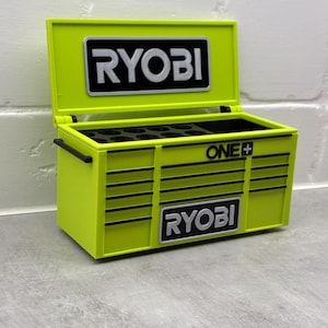 Może przedstawiać: Limonkowo-zielona skrzynka narzędziowa z logo "RYOBI" w kolorze czarno-białym z przodu i na g&oacute;rze. Skrzynia jest otwarta, ukazując przegr&oacute;dki wewnątrz. Widoczne jest r&oacute;wnież logo "ONE+".