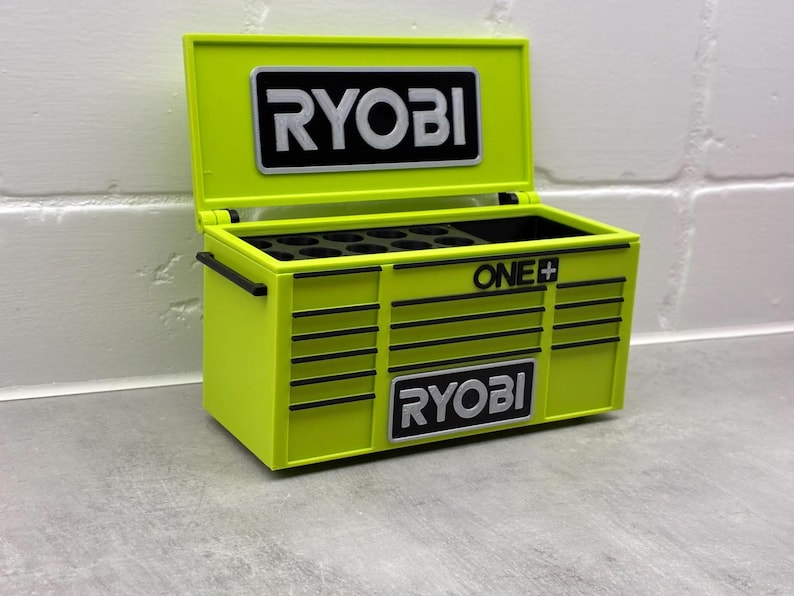 Może przedstawiać: Limonkowo-zielona skrzynka narzędziowa z logo "RYOBI" w kolorze czarno-białym z przodu i na g&oacute;rze. Skrzynia jest otwarta, ukazując przegr&oacute;dki wewnątrz. Widoczne jest r&oacute;wnież logo "ONE+".