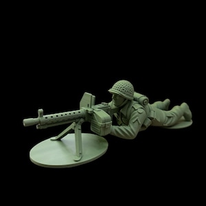 May include: A green plastic toy soldier in a prone firing position, operating a machine gun. The soldier wears a helmet and uniform, with a backpack. The machine gun is mounted on a circular base, and the background is black.