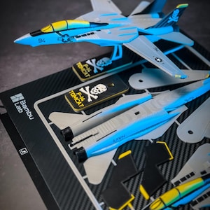 Może przedstawiać: Zestaw modelu samolotu F-14 Tomcat w kolorze niebieskim, szarym i żółtym. Zestaw zawiera niezmontowane części i pudełko z napisem "Bambu Lab" i "F-14 TOMCAT". Samolot ma numer "94" z boku.