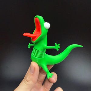 Könnte beinhalten: Eine leuchtend grüne Echsenfigur mit weit geöffnetem Maul, einer langen roten Zunge und einem einzelnen großen weißen Auge. Die Echse hat einen texturierten, segmentierten Mittelteil und einen langen, geschwungenen Schwanz mit kleinen Gliedmaßen.