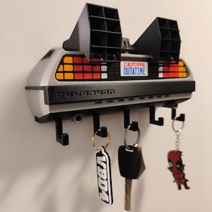 Puede incluir: Un llavero DeLorean gris y negro montado en una pared blanca. El llavero tiene una placa de matrícula que dice "OUTATIME" e incluye llaveros con varios diseños. El llavero tiene cuatro ganchos.