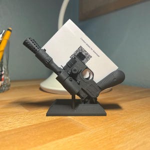 May include: A black 3D printed model of a blaster pistol, designed to hold business cards. The model is on a small black base and has a card holder on the side. The card holder has a white card with the text "Contact details for download" printed on it.