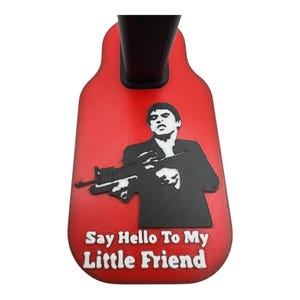 Op de afbeelding: Rode plasticen standaard met een zwart silhouet van een man die een pistool vasthoudt. De standaard heeft de tekst "Say Hello To My Little Friend" in wit.