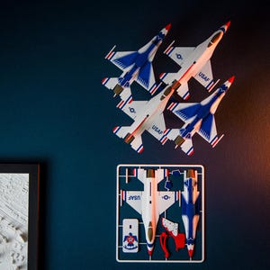 Puede incluir: Tres modelos de aviones de combate con detalles blancos, azules y rojos están montados en una pared azul oscuro. Los aviones están etiquetados como "USAF". Debajo, se ve un kit de modelo de un avión similar, con piezas sin montar.
