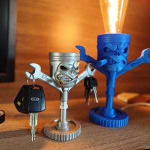 Könnte beinhalten: Zwei schädelartige Lampenhalter, einer silberfarben und einer blau, jeweils mit einem Schraubenschlüsselarm. Der silberne Lampenhalter hat einen Schlüssel befestigt. Der blaue Lampenhalter hat eine Glühbirne. Die Schraubenschlüssel sind mit "17mm" markiert.