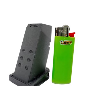 May include: A black 9mm magazine extension for a pistol, next to a green Bic lighter for size comparison.