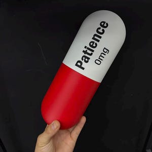 May include: A large, cylindrical capsule-shaped object with a smooth finish. The top half is white with the black text "Patience" and "0mg" printed on it, while the bottom half is a solid, vibrant red color. The object is held in a hand.