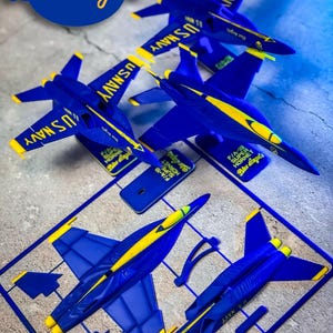 Peut inclure: Maquettes d'avions bleus et jaunes avec le texte "US NAVY" et les mots "Blue Angels". L'image montre des kits de modèles assemblés et non assemblés d'avions F/A-18 Super Hornet.