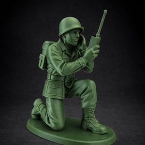May include: A green plastic toy soldier in a kneeling position, holding a radio. The figure wears a helmet, uniform, and boots. The toy is on a green oval base, with a black background.