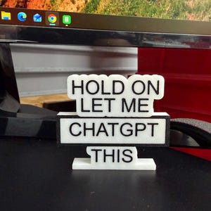 May include: A white and black sign that reads "HOLD ON LET ME CHATGPT THIS." The sign is made of a 3D printed material and is displayed on a black surface. The background includes a computer monitor.