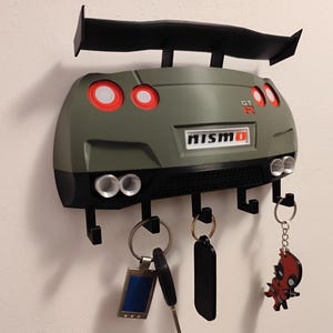 Puede incluir: Un llavero con forma de coche verde y negro con alerón, luces traseras y tubos de escape. El llavero tiene la palabra "NISMO" y tiene ganchos para llaves. Varias llaves y llaveros cuelgan de los ganchos.