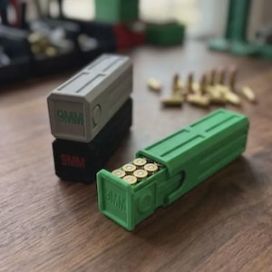 Puede incluir: Tres cajas rectangulares de plástico para almacenamiento de munición con tapas deslizantes. Una caja verde está abierta, revelando ocho cartuchos de latón de 9 mm en su interior. Dos cajas cerradas están apiladas, una gris con texto 9MM verde y una negra con texto 9MM rojo.
