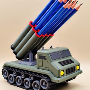 Pode incluir: Um porta-lápis de novidade em forma de veículo militar. A base verde-oliva assemelha-se a um tanque, suportando vários tubos cinzentos com bordas vermelhas, cada um contendo um lápis azul. Os lápis estão afiados. Faróis amarelos e esteiras pretas completam o design.