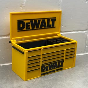 Może przedstawiać: Ż&oacute;łta skrzynka narzędziowa z czarnym logo "DeWALT" na pokrywie i przodzie. Skrzynka jest otwarta, ukazując czarne wnętrze z przegr&oacute;dkami. Słowa "GUARANTEED TOUGH" znajdują się pod logo. Skrzynka ma czarne uchwyty.