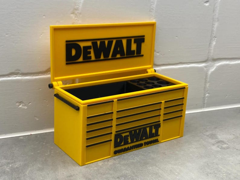 Może przedstawiać: Ż&oacute;łta skrzynka narzędziowa z czarnym logo "DeWALT" na pokrywie i przodzie. Skrzynka jest otwarta, ukazując czarne wnętrze z przegr&oacute;dkami. Słowa "GUARANTEED TOUGH" znajdują się pod logo. Skrzynka ma czarne uchwyty.