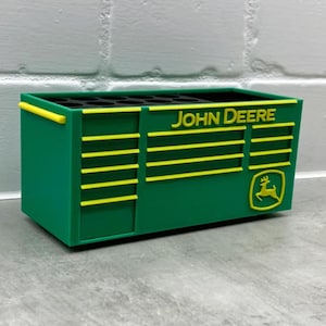 Może przedstawiać: Zielone i ż&oacute;łte prostokątne pudełko do przechowywania z napisem "JOHN DEERE" i logo. Pudełko ma ż&oacute;łte poziome paski i ż&oacute;łty uchwyt. Pudełko stoi na szarej powierzchni.